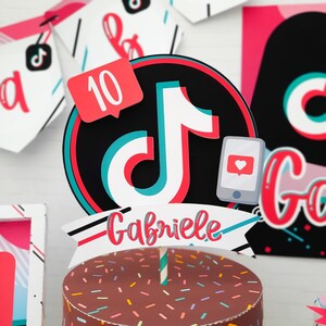 May include: A cake topper featuring the TikTok logo in black, red, and teal, with the name "Gabriele" in red script. Includes a red notification bubble with the number 10 and a smartphone with a heart icon. The cake is brown with colorful sprinkles.