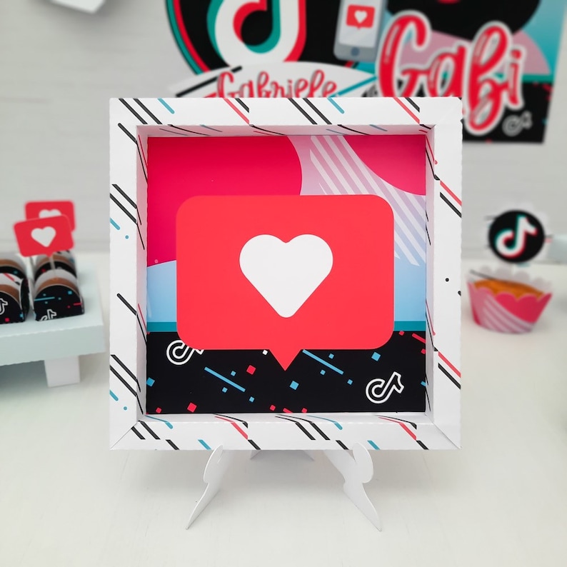 May include: A white framed decoration featuring a red speech bubble with a white heart, a black and white background, and the TikTok logo. The frame has a white background with black, red, and blue diagonal lines. The name "Gabriele" is visible in the background.