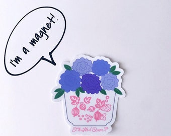 May include: A white ceramic pot with blue and purple flowers and pink berries. The pot has a pink and white floral design. The sticker says "I'm a magnet!" and "Thrifted Queen".