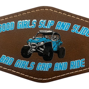 May include: Brown leather patch with stitched edges featuring a blue off-road vehicle graphic. The text "GOOD GIRLS SLIP AND SLIDE" and "BAD GIRLS GRIP AND RIDE" are in blue.