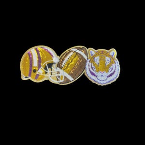May include: Three beaded items on a black background: a football helmet, a football, and a tiger head. The helmet is gold with purple stripes. The football is brown and white. The tiger head is gold, purple, and white.