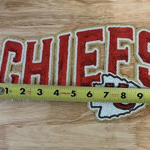 May include: A beaded Chiefs logo, approximately 9 inches long, featuring red lettering outlined in white and gold. The logo includes the team's arrowhead symbol. The item is displayed on a wooden surface.