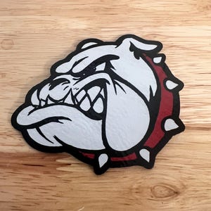 May include: A white and black bulldog head with a red collar, set against a wooden background. The bulldog has an aggressive expression with visible teeth. The collar has white spikes. The image is a graphic design.