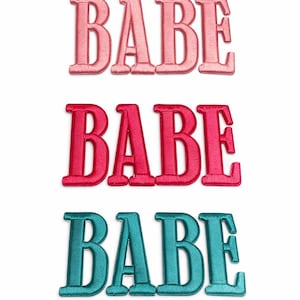 May include: Three embroidered patches spelling the word "BABE" in different colors. The top patch is light pink, the middle is red, and the bottom is teal. The patches are on a white background.