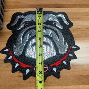 May include: A black sequined bulldog patch, approximately 9 inches tall, with silver and red accents. The patch features a red collar with white spikes and red eyes. The bulldog's face is detailed with silver sequins.