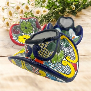 以下が含まれることがあります： 青、黄色、赤、緑の花柄が施されたカラフルなセラミック製のカウボーイハット2つ。伝統的なメキシコのタラベラ様式で作られています。