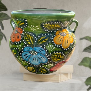 May include: A hand-painted ceramic pot with a green exterior and floral designs in orange, blue, and yellow. The pot has two handles and is supported by a wooden stand. The floral design includes white dots and green leaves.