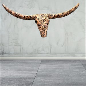 May include: A decorative longhorn skull with a detailed black floral pattern on a tan background. The skull is mounted on a light grey wall, with a grey floor in the foreground.
