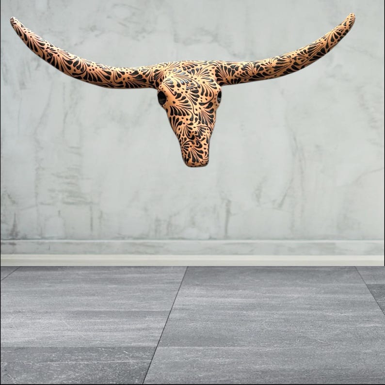 May include: A decorative longhorn skull with a detailed black floral pattern on a tan background. The skull is mounted on a light grey wall, with a grey floor in the foreground.