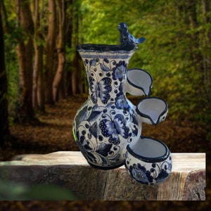 Puede incluir: Un jarr&oacute;n de cer&aacute;mica azul y blanco con una figura de p&aacute;jaro en la parte superior. El jarr&oacute;n presenta un dise&ntilde;o de mariposas y flores. Tiene tres aberturas en forma de pico y un cuenco peque&ntilde;o a juego. El jarr&oacute;n est&aacute; sobre una superficie de madera.