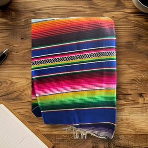 K&ouml;nnte beinhalten: Eine bunte mexikanische Decke mit Streifen in Rot, Orange, Gelb, Gr&uuml;n, Blau und Pink. Die Decke ist gefaltet und hat Fransen an den R&auml;ndern.