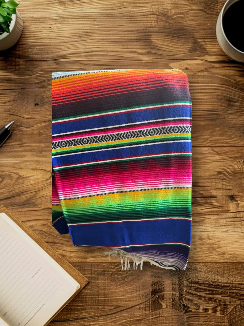 K&ouml;nnte beinhalten: Eine bunte mexikanische Decke mit Streifen in Rot, Orange, Gelb, Gr&uuml;n, Blau und Pink. Die Decke ist gefaltet und hat Fransen an den R&auml;ndern.