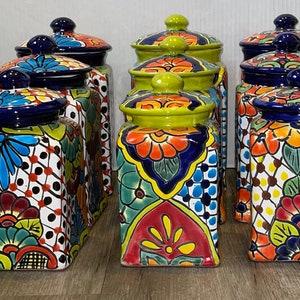 May include: Set of six colorful ceramic canisters with floral designs and blue and green accents. The canisters have square bodies and lids with rounded tops.