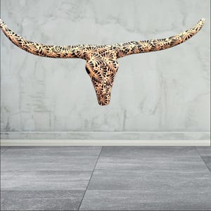 May include: A decorative bull skull wall hanging. The skull is tan with a black floral pattern. The horns are long and curved. The skull is suspended against a grey wall and floor.