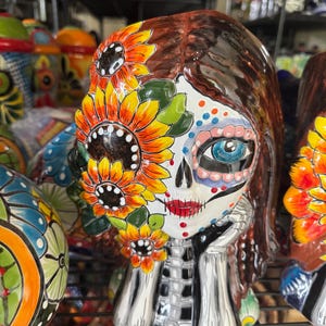 May include: A Day of the Dead ceramic figurine with a painted skull face, brown hair, and a sunflower design. The figurine is resting its chin on its hand. Other colorful ceramic pieces are visible in the background.