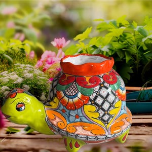 Puede incluir: Una maceta de cerámica con forma de tortuga, con un diseño pintado a mano. El caparazón de la tortuga presenta un colorido patrón floral con detalles en rojo, naranja, azul y verde. La maceta tiene un borde naranja y un interior blanco.