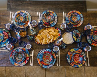 May include: A table setting featuring colourful, hand-painted ceramic plates, bowls, and mugs. The plates have intricate floral designs in shades of blue, green, yellow, and red. A bowl of tortilla chips is in the centre of the table.
