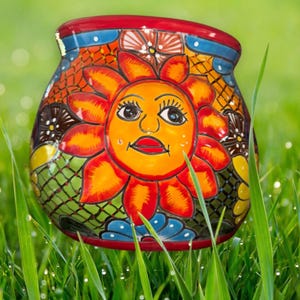 May include: A colourful ceramic planter with a hand-painted design of a smiling sun with a red mouth and blue eyes. The planter is decorated with green, orange, red, blue and yellow flowers and leaves.