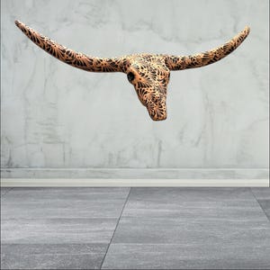 May include: A decorative wall-mounted bull skull with long horns. The skull is tan with intricate black floral patterns. The horns extend outward, creating a striking silhouette. The piece is designed to be a unique home decor item.