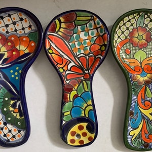 Pode incluir: Três repousos de colher de cerâmica coloridos com desenhos florais intrincados. Os repousos de colher são pintados em tons de azul, verde, vermelho, amarelo e laranja. Cada repouso de colher tem um design diferente.