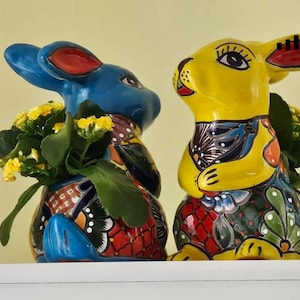 May include: Two ceramic bunny planters, one blue and one yellow, with colorful painted designs. Both planters have green plants with yellow flowers inside.