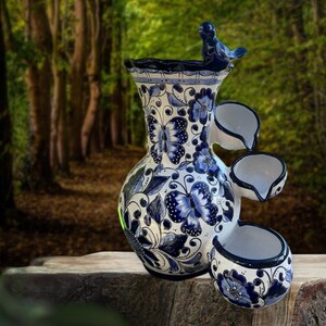 Puede incluir: Una fuente de cer&aacute;mica blanca y azul con un dise&ntilde;o de mariposas y flores. La fuente tiene tres cuencos en cascada y una peque&ntilde;a figura de p&aacute;jaro en la parte superior. La fuente se coloca sobre una superficie de madera con un fondo borroso de bosque.