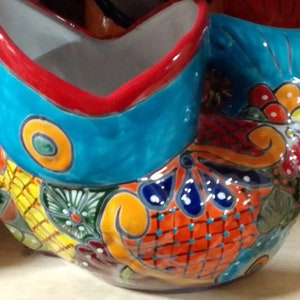 May include: A colorful ceramic planter shaped like a fish with a blue, red, and white design. The planter has a wide opening at the top and a small opening at the bottom for drainage.