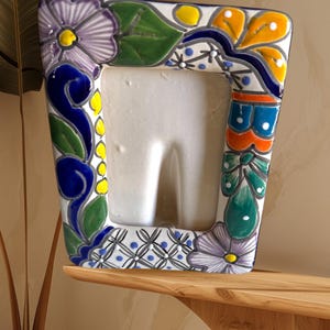 Puede incluir: Un marco de fotos de cerámica blanca con un diseño floral colorido. El marco tiene un patrón floral azul, verde, amarillo y morado alrededor del borde.