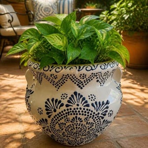 May include: A cream-coloured ceramic planter with two side handles, decorated with dark blue painted patterns including dots, zigzags, and floral motifs. The pot contains a lush green pothos plant with variegated heart-shaped leaves.