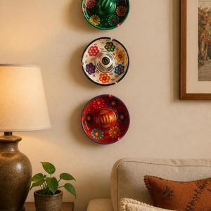 May include: Three circular ceramic ashtrays shaped like miniature sombreros are mounted vertically on a wall. The top one is green, the middle is white with a black rim, and the bottom is red. Each features colorful painted floral patterns.