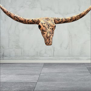 May include: A decorative steer skull with long horns, featuring an intricate black floral pattern on a tan background. The skull is suspended against a neutral grey wall and floor. This piece is ideal for rustic or southwestern decor.