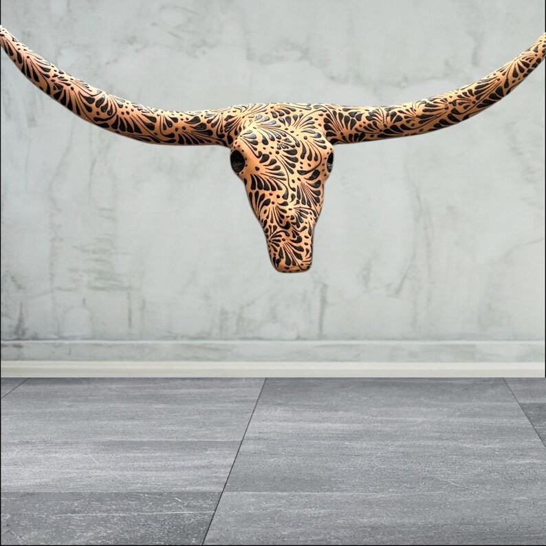 May include: A decorative steer skull with long horns, featuring an intricate black floral pattern on a tan background. The skull is suspended against a neutral grey wall and floor. This piece is ideal for rustic or southwestern decor.