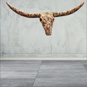 May include: A decorative longhorn skull featuring a detailed black floral pattern on a tan base. The skull is mounted on a light grey wall, creating a striking contrast. The horns extend outwards, adding to the piece's visual impact.