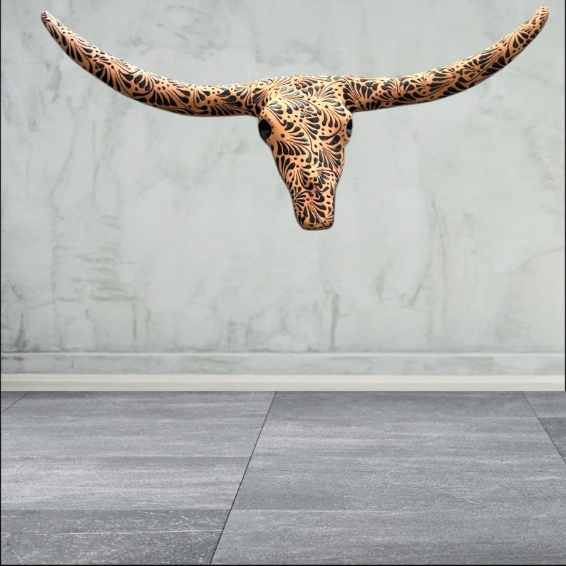 May include: A decorative longhorn skull featuring a detailed black floral pattern on a tan base. The skull is mounted on a light grey wall, creating a striking contrast. The horns extend outwards, adding to the piece's visual impact.