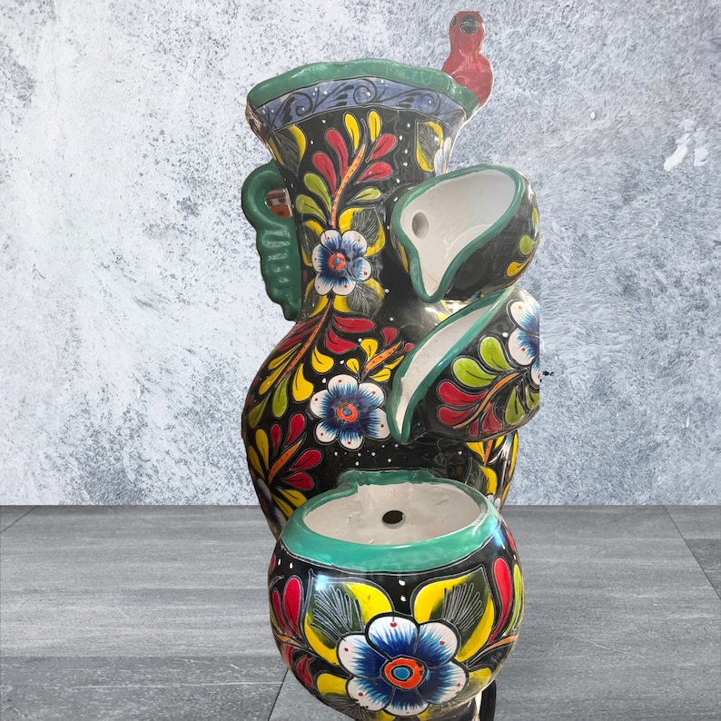 Puede incluir: Una fuente alta de cer&aacute;mica negra con un vibrante dise&ntilde;o floral en rojo, amarillo y azul. La fuente tiene m&uacute;ltiples niveles y un peque&ntilde;o p&aacute;jaro rojo en la parte superior. Los bordes son verdes.