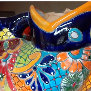 May include: A colorful ceramic fish planter with blue, orange, yellow, green, and red floral designs. The planter has a wide opening at the top for planting.