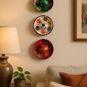 May include: Three circular ceramic ashtrays shaped like miniature sombreros are mounted vertically on a wall. The top one is green, the middle is white with a black rim, and the bottom is red. Each features colorful painted floral patterns.