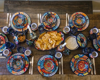 May include: A table setting featuring colourful, hand-painted ceramic dinnerware, bowls, and mugs. The plates have intricate floral designs in shades of blue, green, yellow, and red. A bowl of tortilla chips is in the centre of the table.