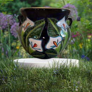 Op de afbeelding: Een donkerblauwe keramische plantenpot met twee handvatten aan de zijkant, voorzien van een handgeschilderd ontwerp van witte calla lelies met oranje harten en groene bladeren. De ronde pot heeft een glanzende afwerking op een witte sokkel.