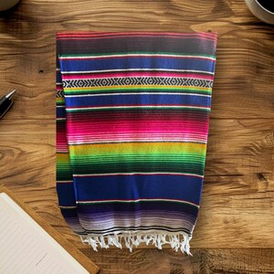 K&ouml;nnte beinhalten: Eine bunte mexikanische Decke mit Streifen in Rot, Rosa, Gr&uuml;n, Blau, Lila und Wei&szlig;. Die Decke hat einen Fransenrand und ist in der Mitte gefaltet.