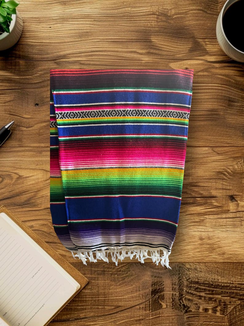K&ouml;nnte beinhalten: Eine bunte mexikanische Decke mit Streifen in Rot, Rosa, Gr&uuml;n, Blau, Lila und Wei&szlig;. Die Decke hat einen Fransenrand und ist in der Mitte gefaltet.