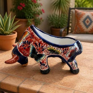 Puede incluir: Una maceta de cerámica con forma de toro que presenta un acabado brillante con una base roja y blanca. La superficie está decorada con motivos de mariposas coloridas en tonos azul, verde y amarillo, con un borde azul oscuro.