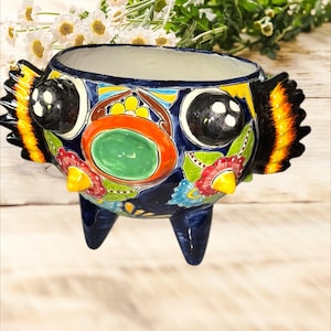Könnte beinhalten: Ein farbenfroher Keramiktopf in Form eines Vogels mit großen Augen, einem grünen Kreis in der Mitte und gelben, orangefarbenen, roten, blauen und schwarzen Details. Der Topf hat drei Beine und ist mit Blumen verziert.