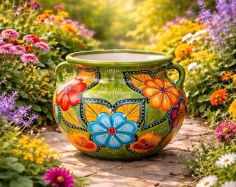 May include: A round, green ceramic planter featuring two side handles and a hand-painted floral pattern with large blue, orange, and red flowers surrounded by yellow leaves and black outlines, set against a vibrant, sunlit garden background.
