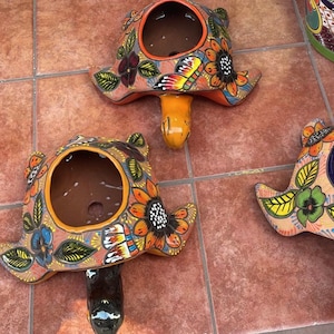 Może przedstawiać: Dwa kolorowe ceramiczne doniczki w kształcie żółwia z namalowanymi kwiatami i motylami. Doniczki są pomarańczowe, brązowe, zielone, żółte, niebieskie i czarne.