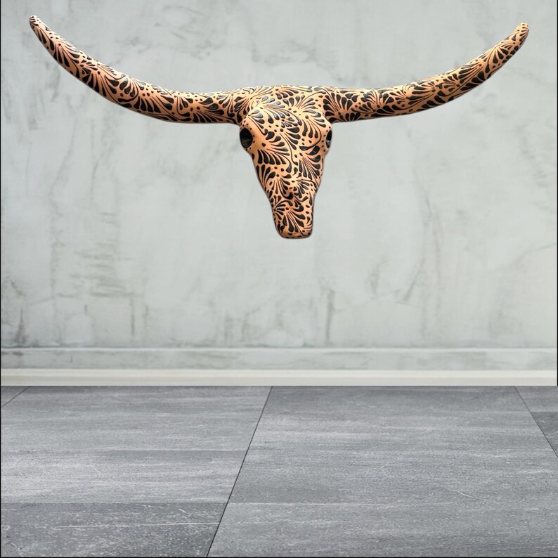 May include: A decorative steer skull with long horns, featuring a detailed black floral pattern on a tan background. The skull is mounted on a light grey wall, creating a rustic and artistic aesthetic.