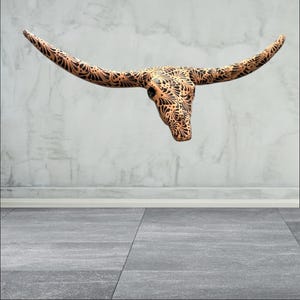 May include: A decorative steer skull with long horns, featuring an intricate black floral pattern on a tan background. The skull is mounted on a wall, showcasing a rustic aesthetic. The horns extend outward, adding to the piece's dramatic appeal.