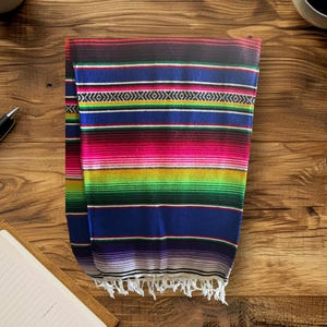 K&ouml;nnte beinhalten: Eine bunte mexikanische Decke mit Streifen in Rot, Rosa, Gr&uuml;n, Blau, Gelb und Wei&szlig;. Die Decke hat einen Fransenrand.