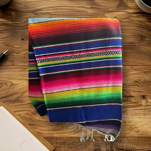 K&ouml;nnte beinhalten: Eine bunte mexikanische Decke mit roten, orangen, gelben, gr&uuml;nen, blauen und schwarzen Streifen. Die Decke ist gefaltet und hat Fransen am unteren Rand.