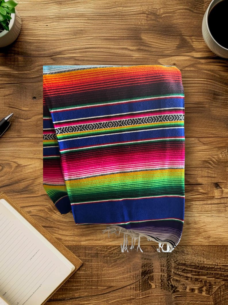 K&ouml;nnte beinhalten: Eine bunte mexikanische Decke mit roten, orangen, gelben, gr&uuml;nen, blauen und schwarzen Streifen. Die Decke ist gefaltet und hat Fransen am unteren Rand.